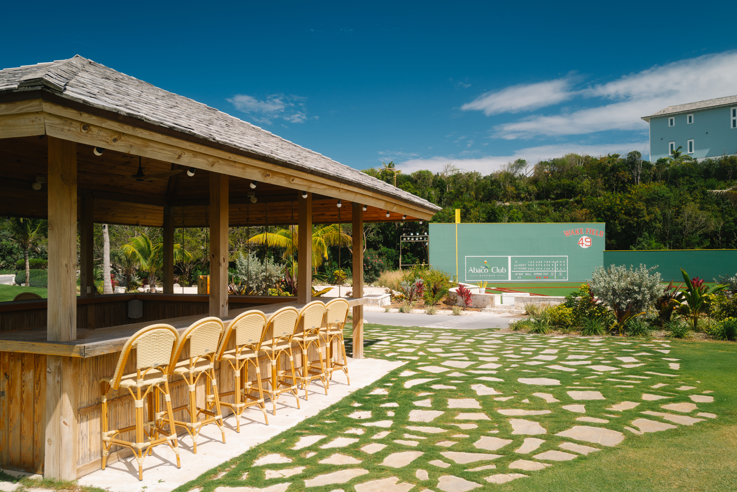 Open-air wooden bar with beige wicker stools under a thatched roof, facing a green baseball-field wall with Wake Field 49.