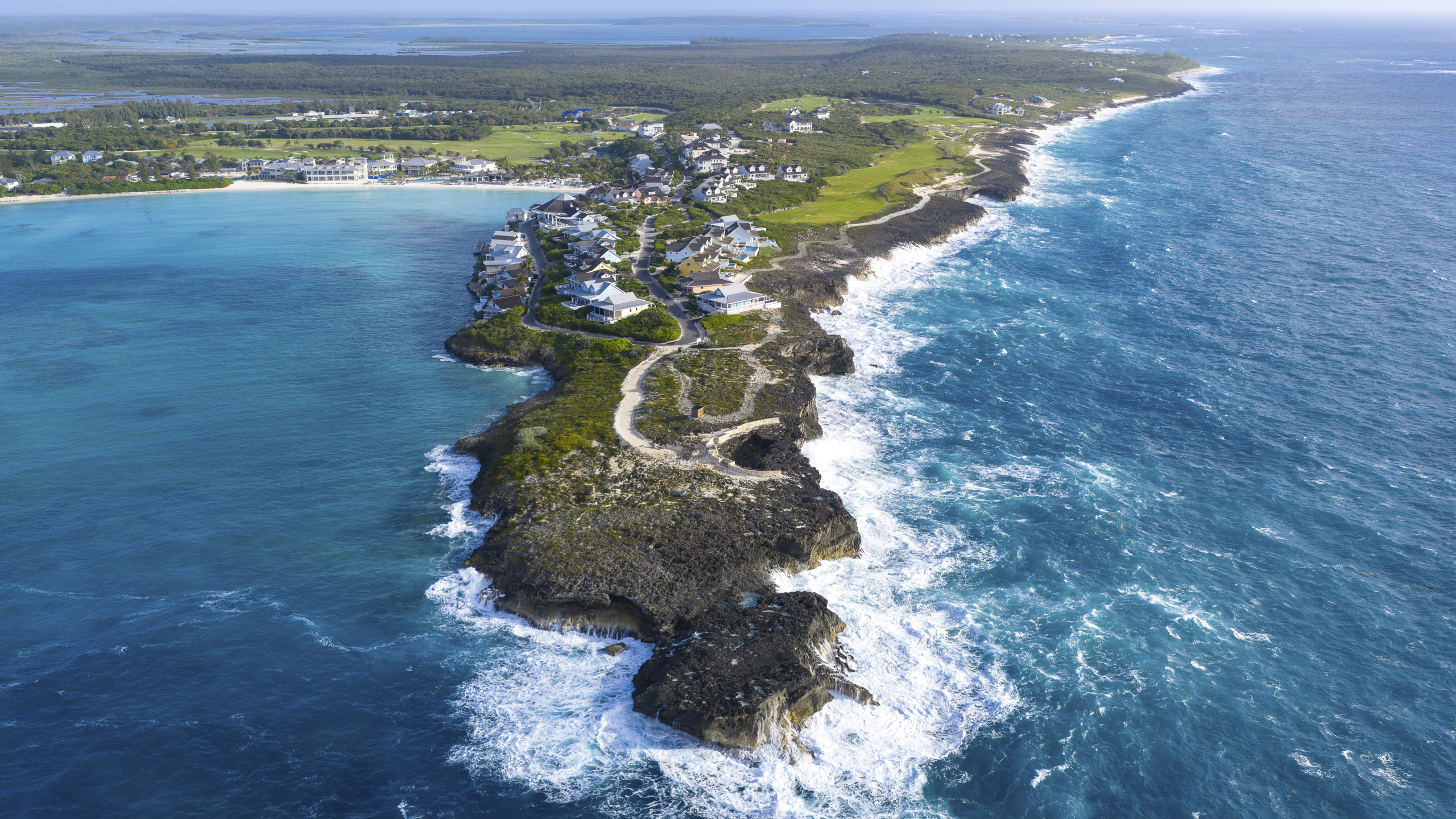 Aerial view of a small coastal island with white houses along a curved shoreline, green lawns, and bright blue ocean.