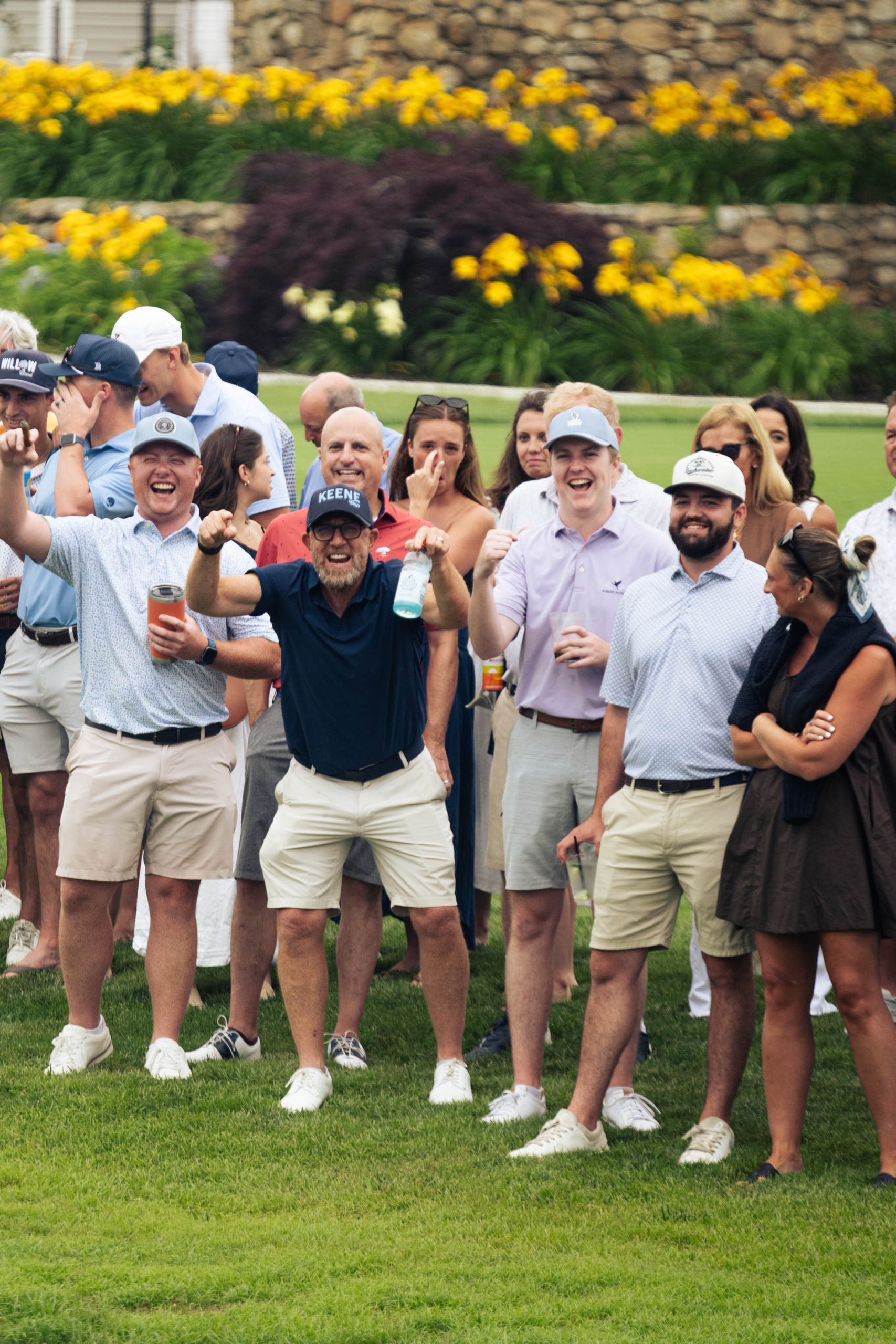 Group of people on a lawn, cheering and posing for a photo at an outdoor event, with yellow flowers and a stone wall in the background.